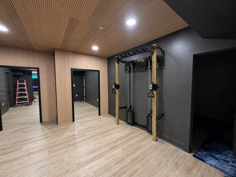 Finished basement gym with a wood slat ceiling and wall, light wood look plank flooring, and a wall mounted rack with cable pulleys on a black wall