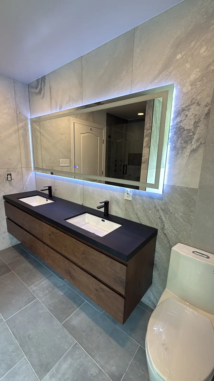 Floating walnut double vanity with a black countertop and matte black faucets under a backlit mirror