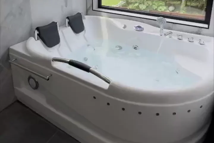 Corner jetted soaking tub filled with water, with two black headrests and chrome fixtures, below a black framed window, with grey tile on the wall and floor