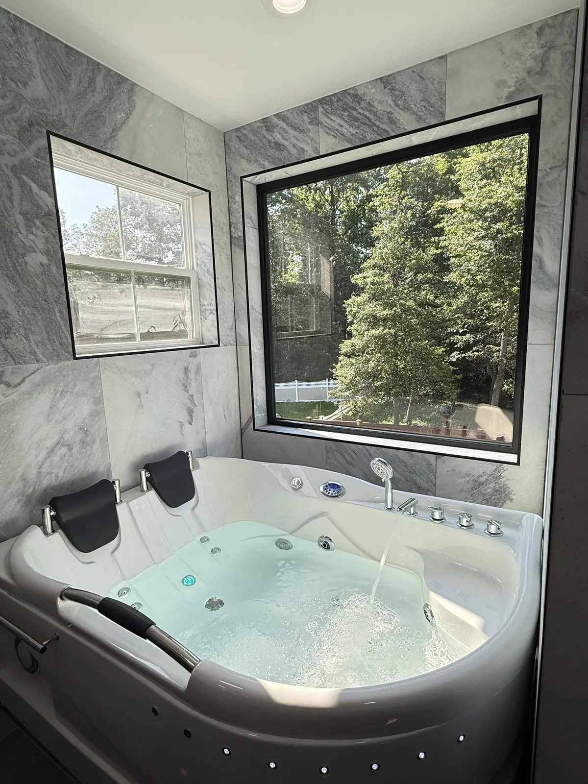 Jetted soaking tub with two black headrests under a large black framed window, surrounded by grey marble look wall tile